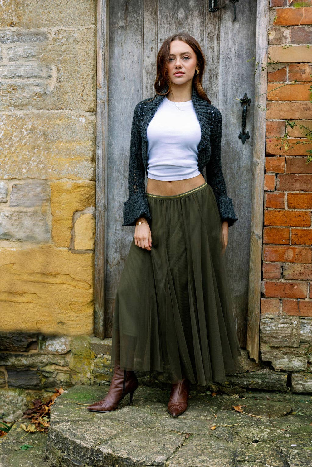 Woman standing in front of a rustic stone wall wearing a white top, green skirt, and brown boots.