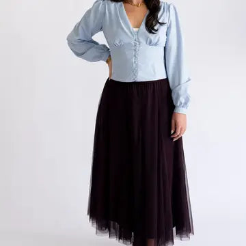 A woman modeling a light blue long-sleeve blouse with ruffle details and a dark brown tulle midi skirt, shown against a plain white studio background.