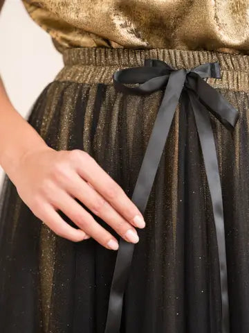 Close-up of a person wearing a gold sequin top and black pleated skirt with a black ribbon, with a blurred background.