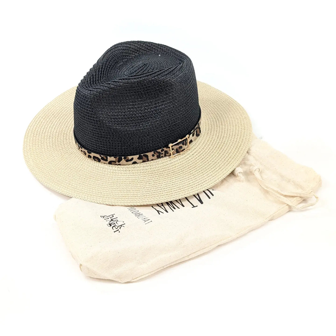 A black woven straw fedora hat with a leopard-print band and a small gold-tone buckle, displayed on top of a cream-colored dust bag with black text.