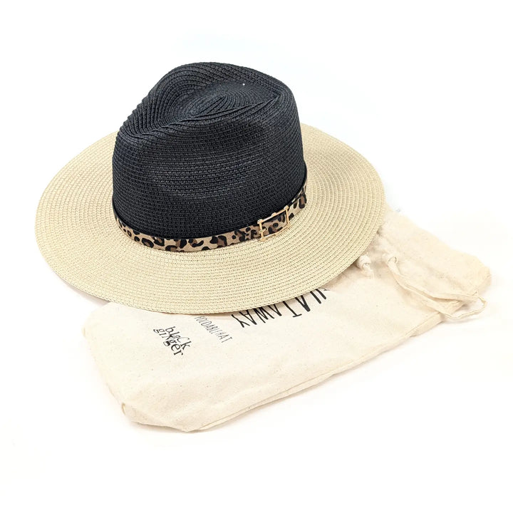 A black woven straw fedora hat with a leopard-print band and a small gold-tone buckle, displayed on top of a cream-colored dust bag with black text.