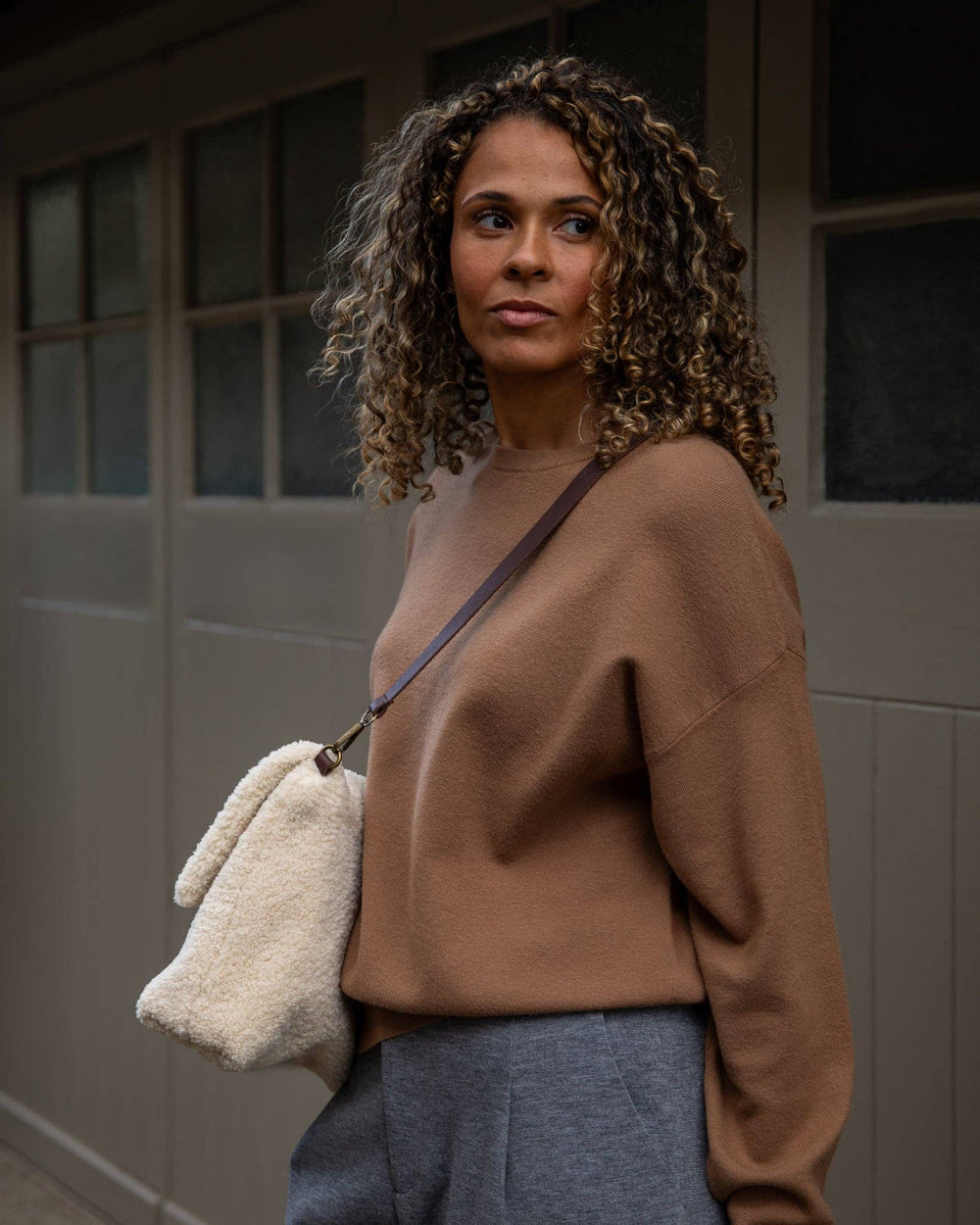 Woman wearing a brown sweater with a white bag over her shoulder, standing against a neutral background.