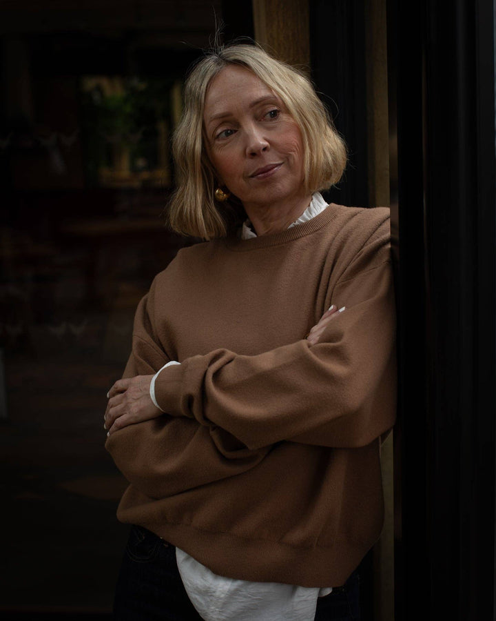 Woman wearing a brown sweater and white shirt in a dimly lit room.