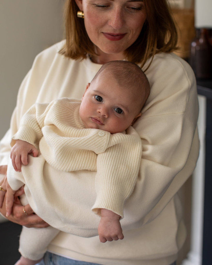 Woman holding a baby wrapped in a cream-colored blanket indoors.