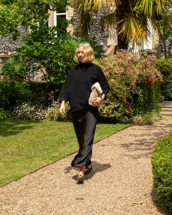 Woman in black outfit walking on a garden path with greenery and a building in the background