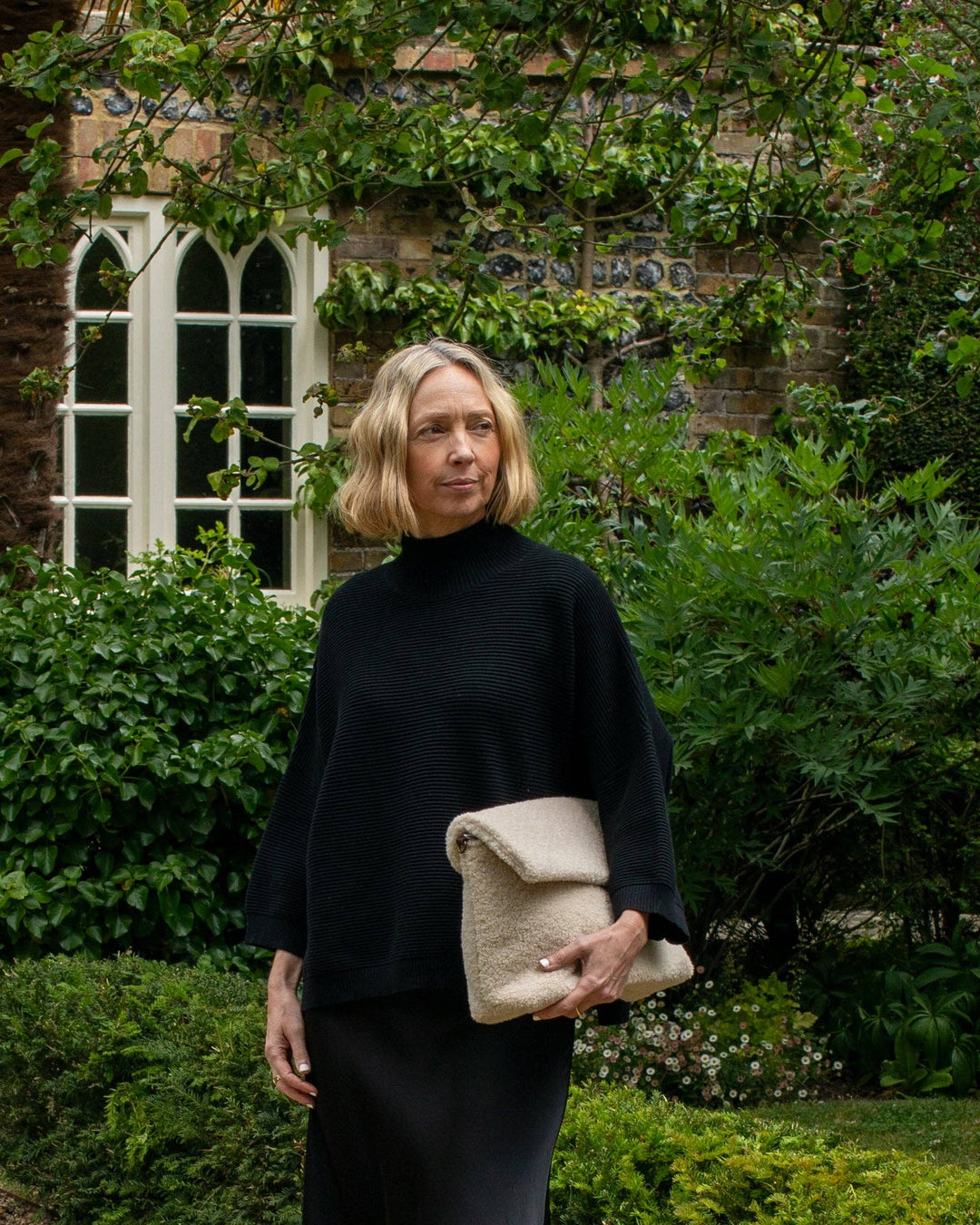 Woman holding a beige clutch in front of a brick building with greenery