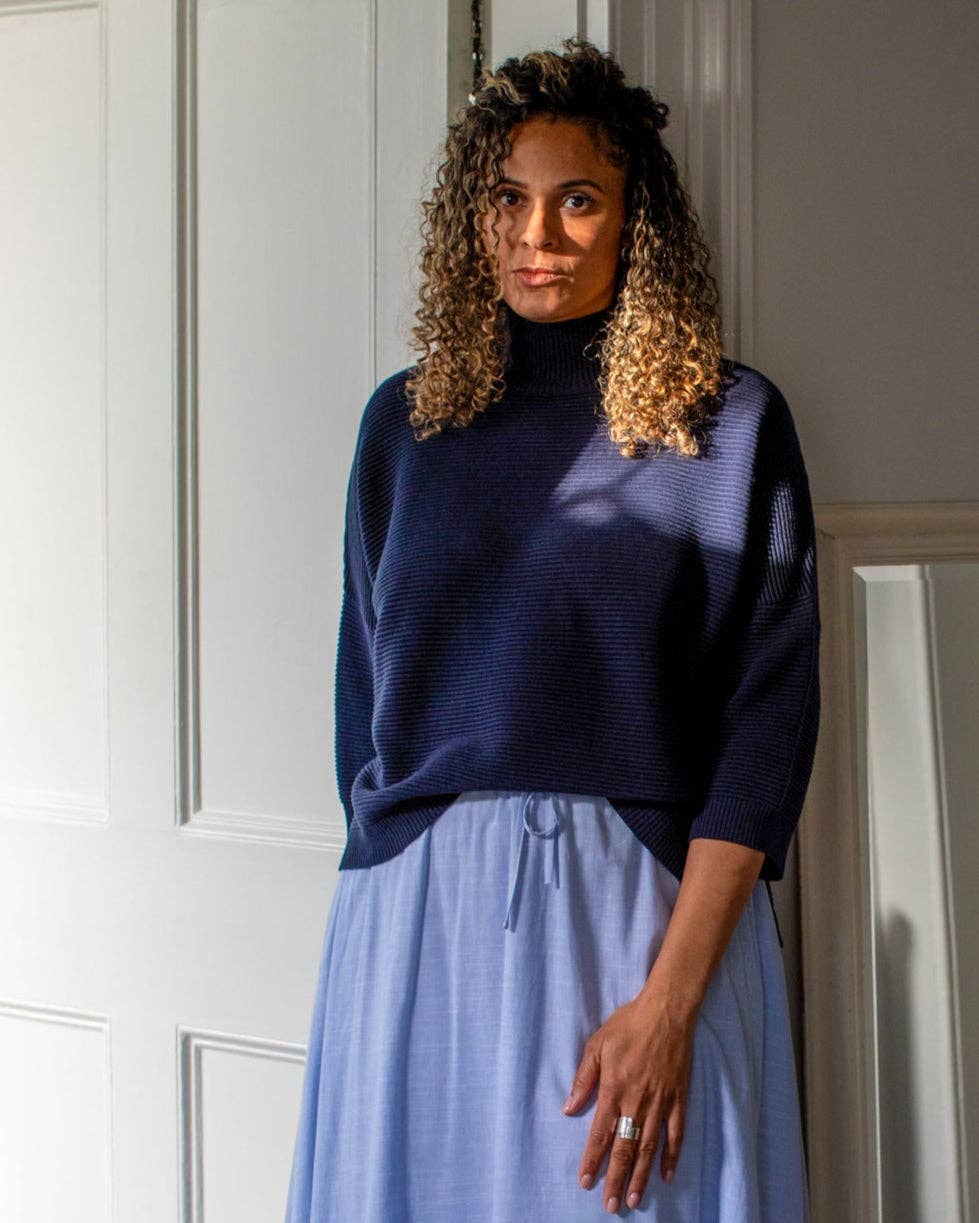 Woman wearing a navy sweater and light blue skirt standing against a white wall.