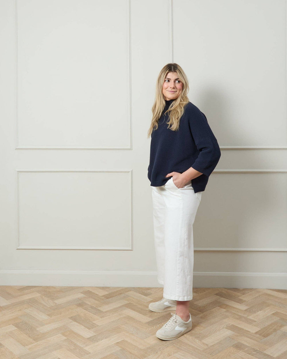 Woman wearing a navy sweater and white pants standing in a room with light-colored walls and wooden floor.