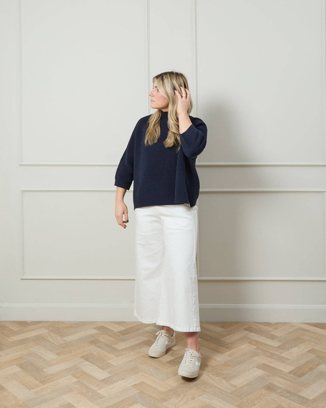 Woman wearing a navy sweater and white skirt standing in a room with wooden flooring and white paneled walls.