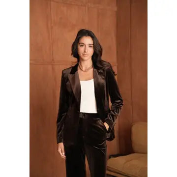 A woman stands indoors wearing a dark brown velvet blazer and matching trousers over a white top, posed against a warm wood-paneled wall with part of a beige chair visible.
