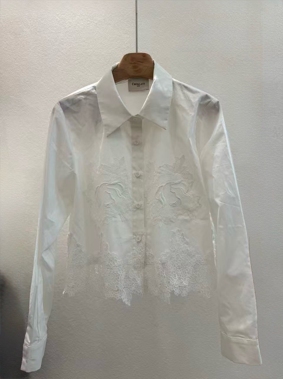 A white long-sleeve button-up shirt with floral embroidery and lace detailing, hanging on a wooden hanger against a textured off-white wall.