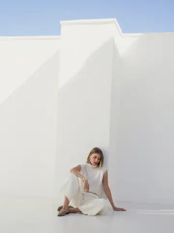 A woman wearing a sleeveless white high-neck dress sits on the floor against a minimalist white architectural wall under a clear blue sky.