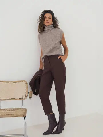 A woman models a sleeveless taupe knit turtleneck sweater and dark brown cropped trousers, standing in a minimalist room with a light-colored wall and a small woven chair.