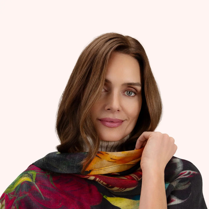 A woman with shoulder-length brown hair models a lightweight scarf with a vibrant floral print in shades of red, yellow, green, and blue over a light beige turtleneck, set against a solid pale pink background.