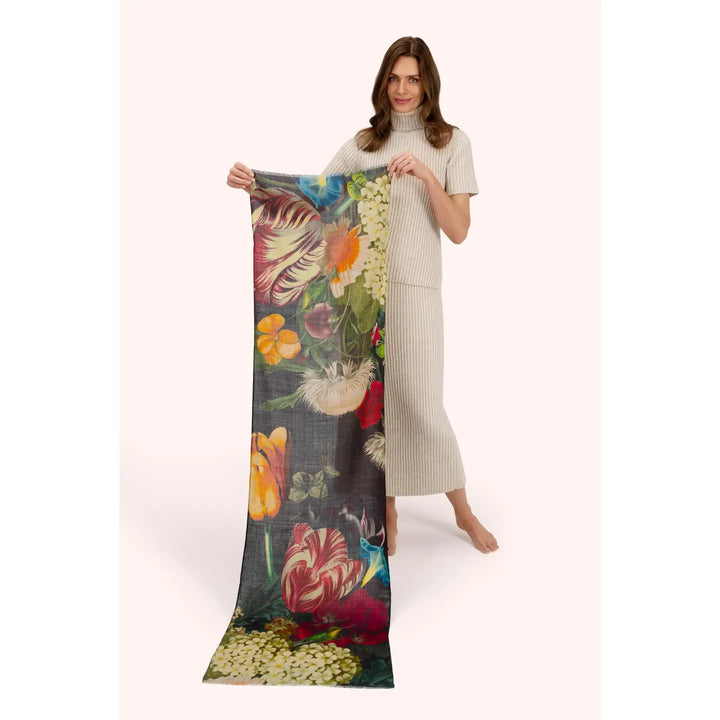 A woman stands barefoot against a pale pink studio background, holding up a long rectangular scarf or fabric panel with a large multicolor floral print. The print includes red, orange, yellow, white, green, and dark blue tones on a dark ground.