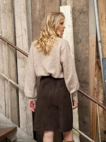 A woman with blonde hair is shown from behind wearing a beige fuzzy knit sweater and a dark brown skirt, standing in front of a rustic wooden wall and staircase.