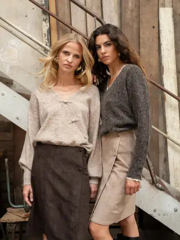 Two women posing together in a rustic indoor setting with wooden walls and metal stairs. The woman on the left wears a light beige knit sweater with toggle details and a dark brown skirt.
