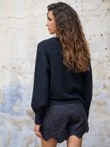 A woman with long, wavy brown hair is shown from behind, wearing a dark navy blue knit sweater and matching dark lace shorts, standing against a textured, light-colored brick wall.