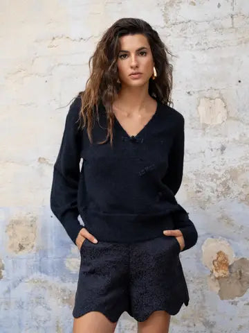 A woman modeling a dark navy blue knit sweater and matching textured shorts against a distressed, light-colored brick wall.