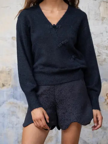 A woman wearing a dark navy textured knit sweater with matching knit shorts, standing against a rustic light gray and beige plaster wall.