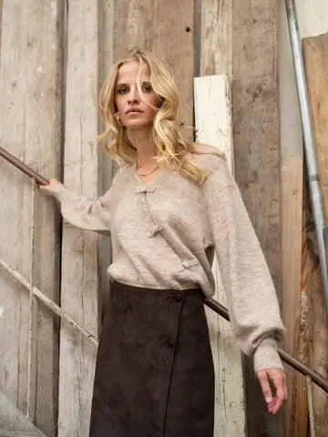 A woman wearing a beige fuzzy long-sleeve top with a split neckline and dark brown skirt, posing in front of a rustic wooden wall.