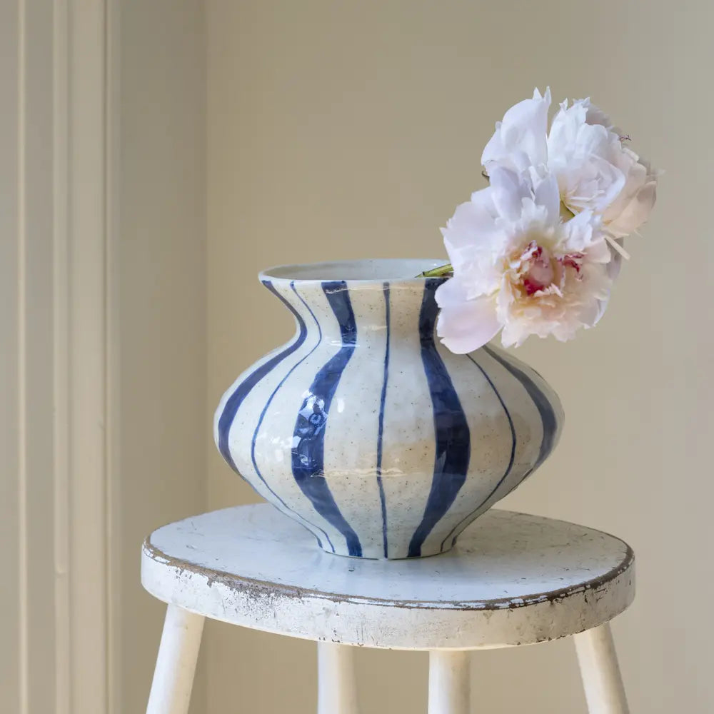 A ceramic vase with a blue and white vertical stripe pattern sits on a distressed white stool, holding a pale pink peony flower against a soft beige wall background.