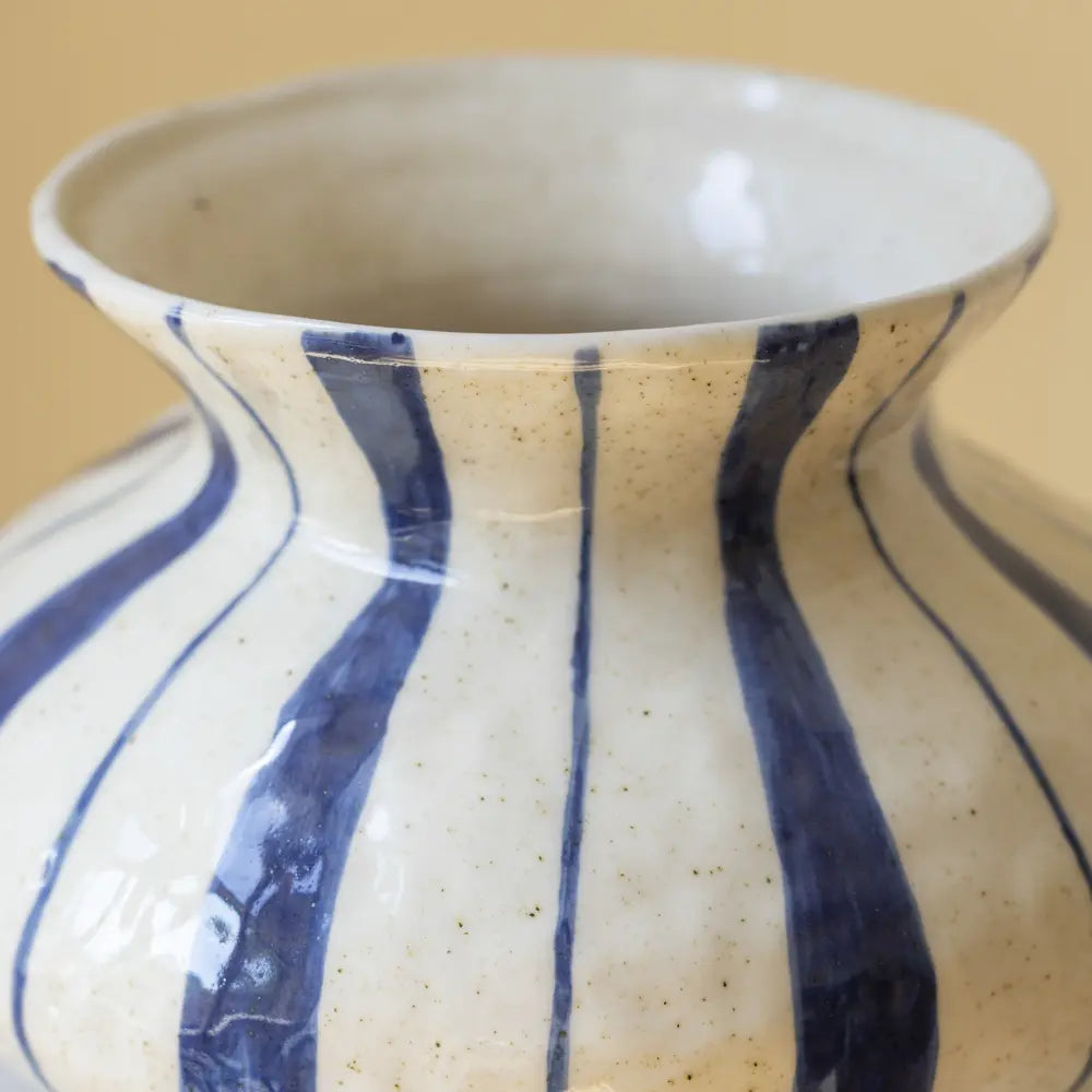 Close-up of a ceramic vase with a cream-colored speckled glaze and bold vertical blue stripes, set against a solid beige background.