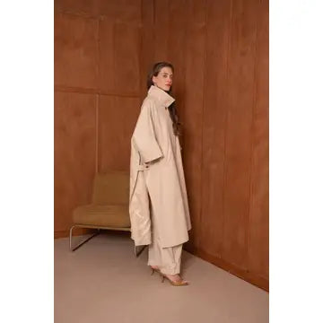 Woman in a beige coat standing against a brown wall.
