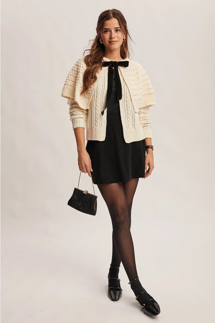 Woman wearing a cream cardigan, black dress, and black shoes on a plain background