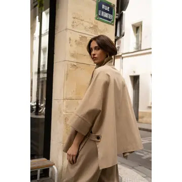 Woman in a beige coat standing on a street corner with a building and street sign in the background.