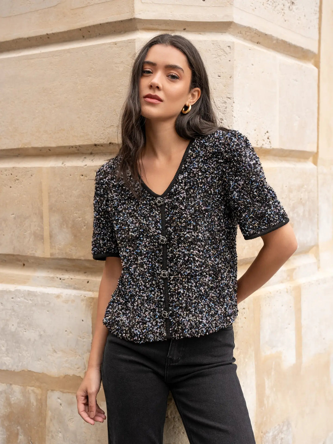 Woman wearing a black floral blouse and black pants standing against a stone wall.